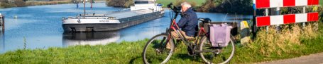 Links ligt de Maas met daarin een groot containerschip. Op de voorgrond een man die zit op een bankje met daarvoor een fiets.